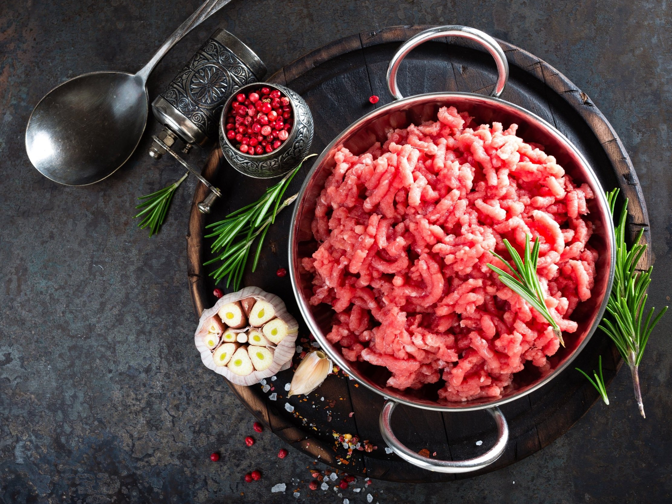 Fresh ground beef in a silver bowl, surrounded by herbs and spices, on a rustic wooden board. Ideal for cooking and meal preparation.