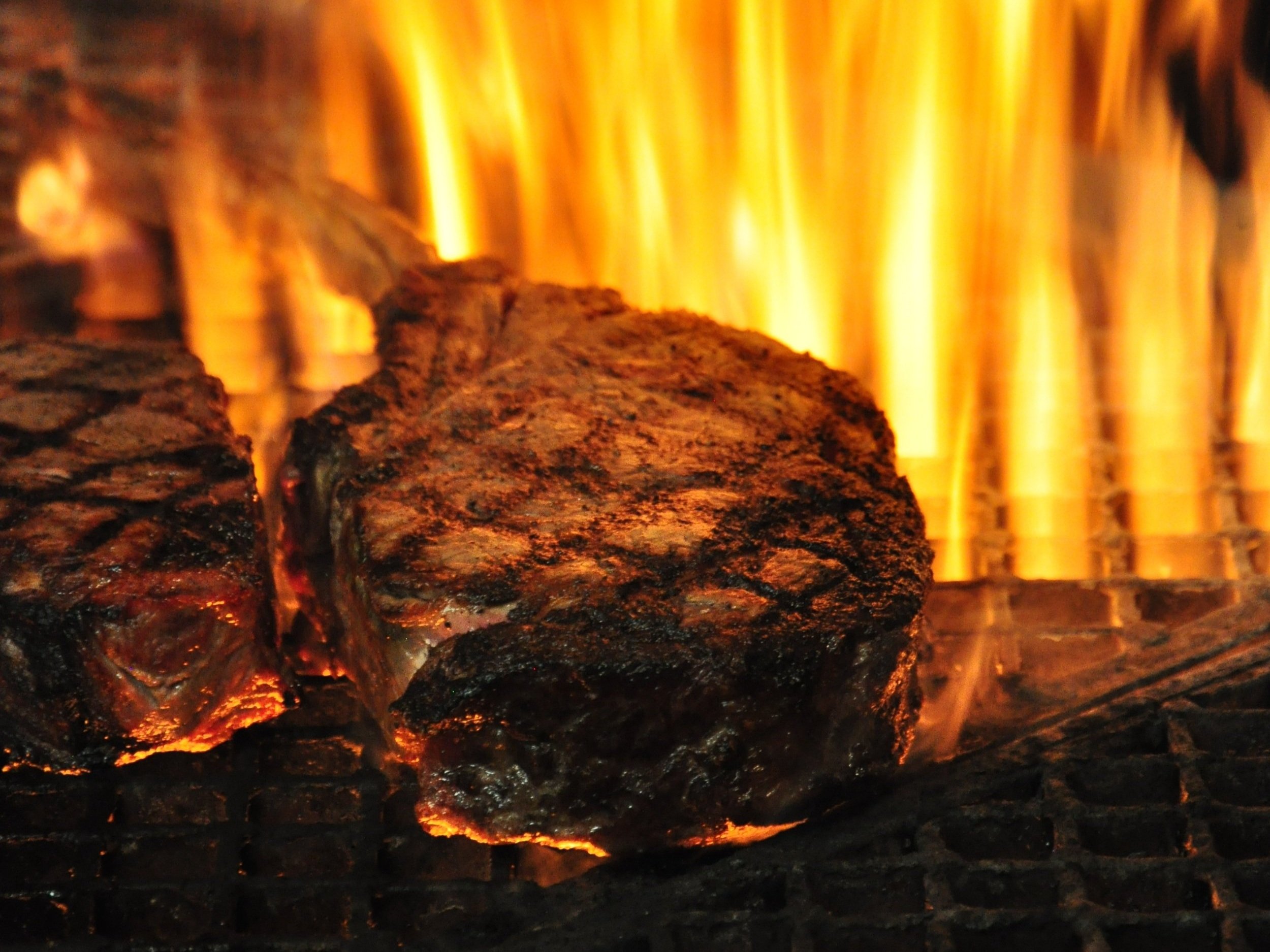 beef_tomahawkribeye_grilled_flame Close-up of two juicy steaks grilling over an intense flame, showcasing charred grill marks and a vibrant fire background, highlighting the art of barbecue cooking.