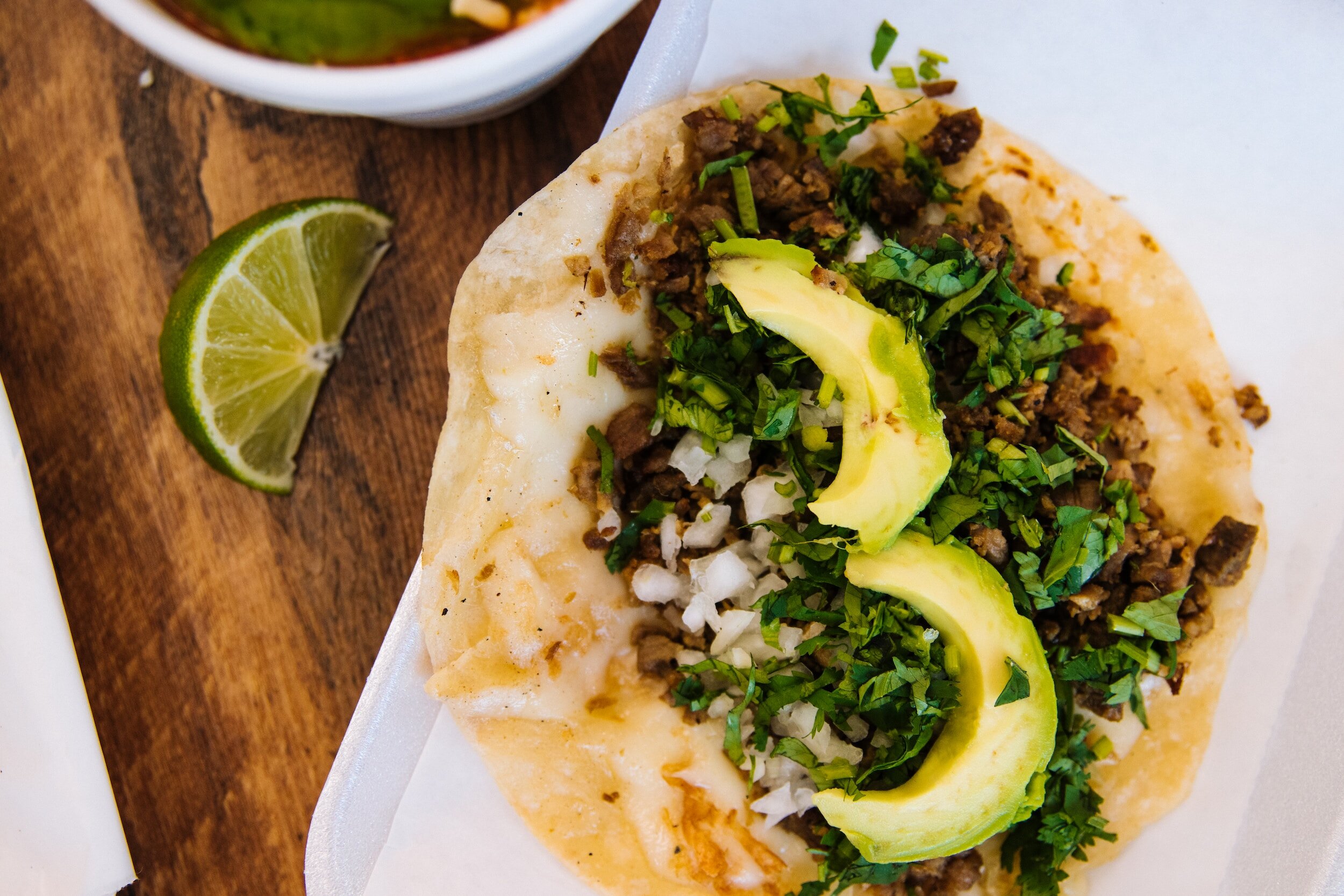 Delicious taco topped with seasoned meat, fresh cilantro, diced onions, and slices of avocado, served with a lime wedge on a wooden table.