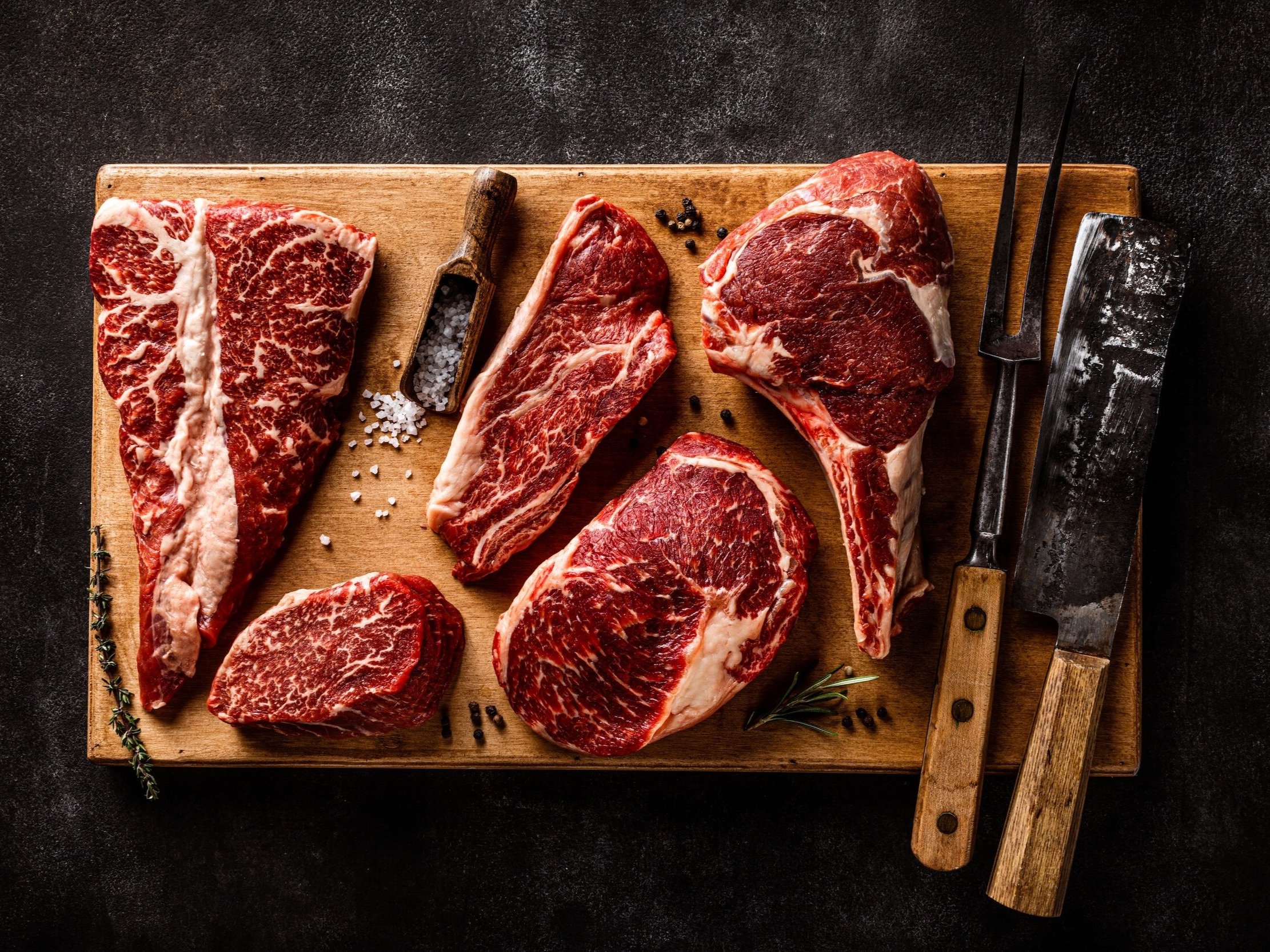 Assorted cuts of marbled beef, including ribeye and sirloin, displayed on a wooden cutting board with salt, herbs, and butchering tools, showcasing high-quality meat for gourmet cooking.