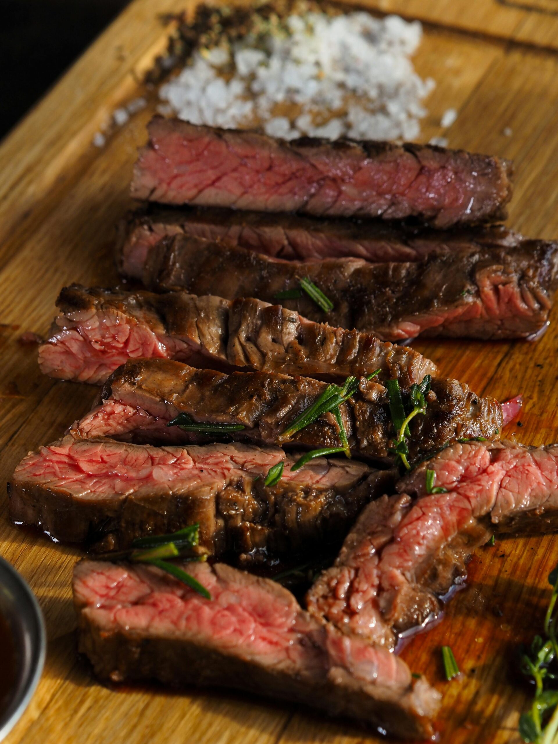 Sliced grilled steak on a wooden cutting board, garnished with fresh herbs and coarse salt, showcasing a juicy, medium-rare interior.